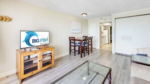 A living room with a television, wooden cabinet, glass coffee table, dining table with chairs, and a door leading to a kitchen.