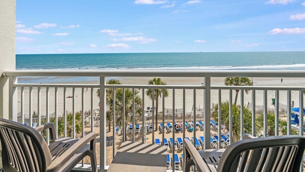 A balcony with a view of the beach, lounge chairs, and palm trees.