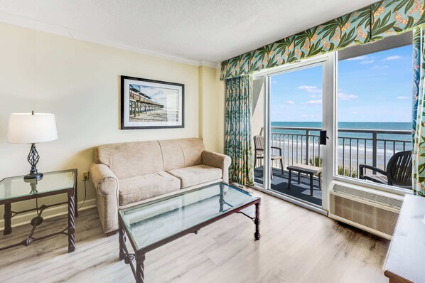 A living room with a sofa, a glass coffee table, a lamp, a framed picture on the wall, and a balcony with a view of the beach.
