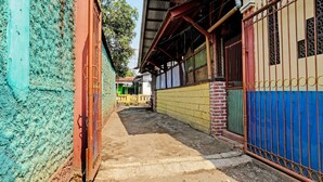 A narrow alleyway with a dirt path, a metal gate, and a building with a corrugated metal roof and colorful walls. - Hotel O Fj Guest House (Cileunyi)