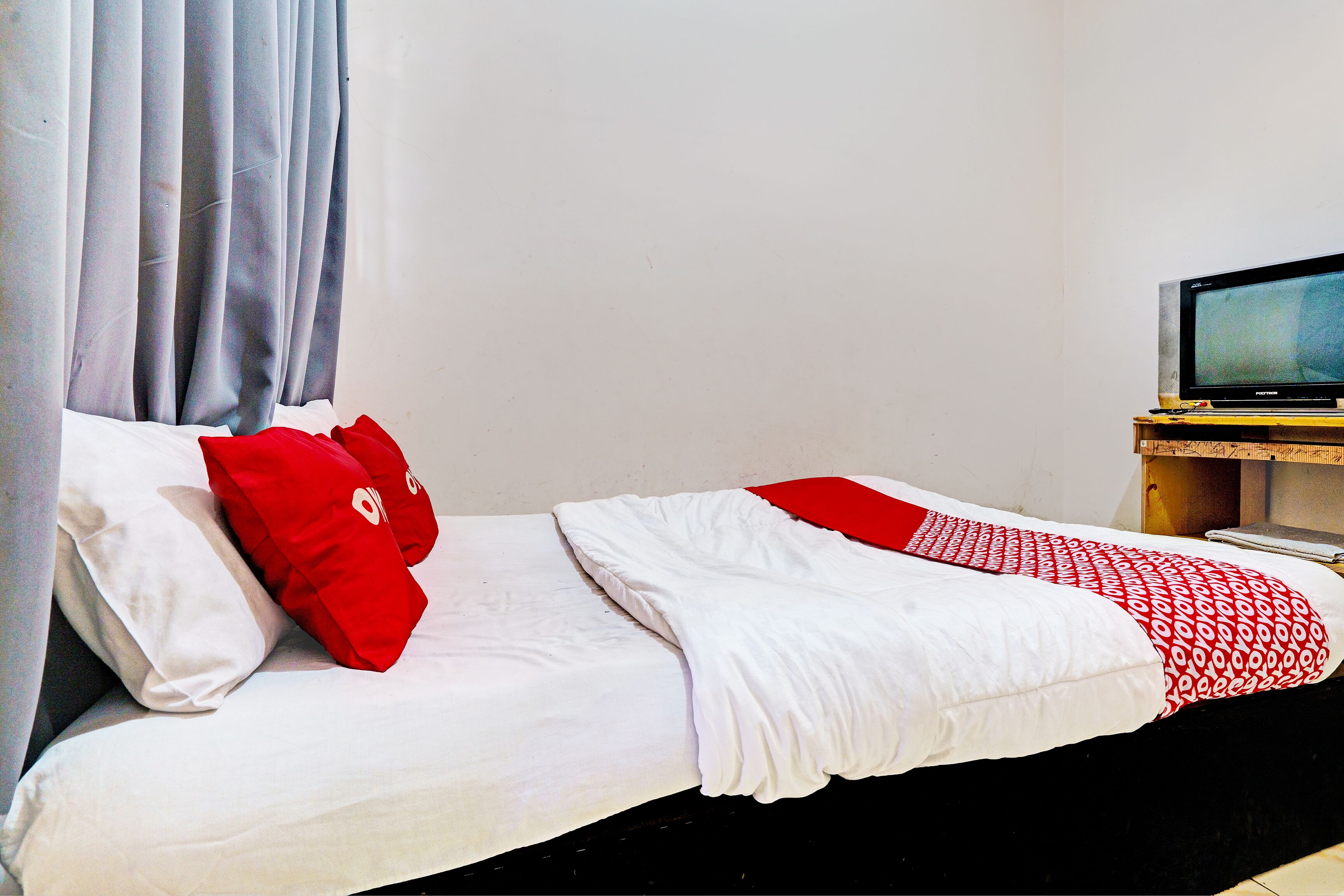 Un lit avec une literie blanche et rouge, un téléviseur posé sur un meuble en bois et un rideau.