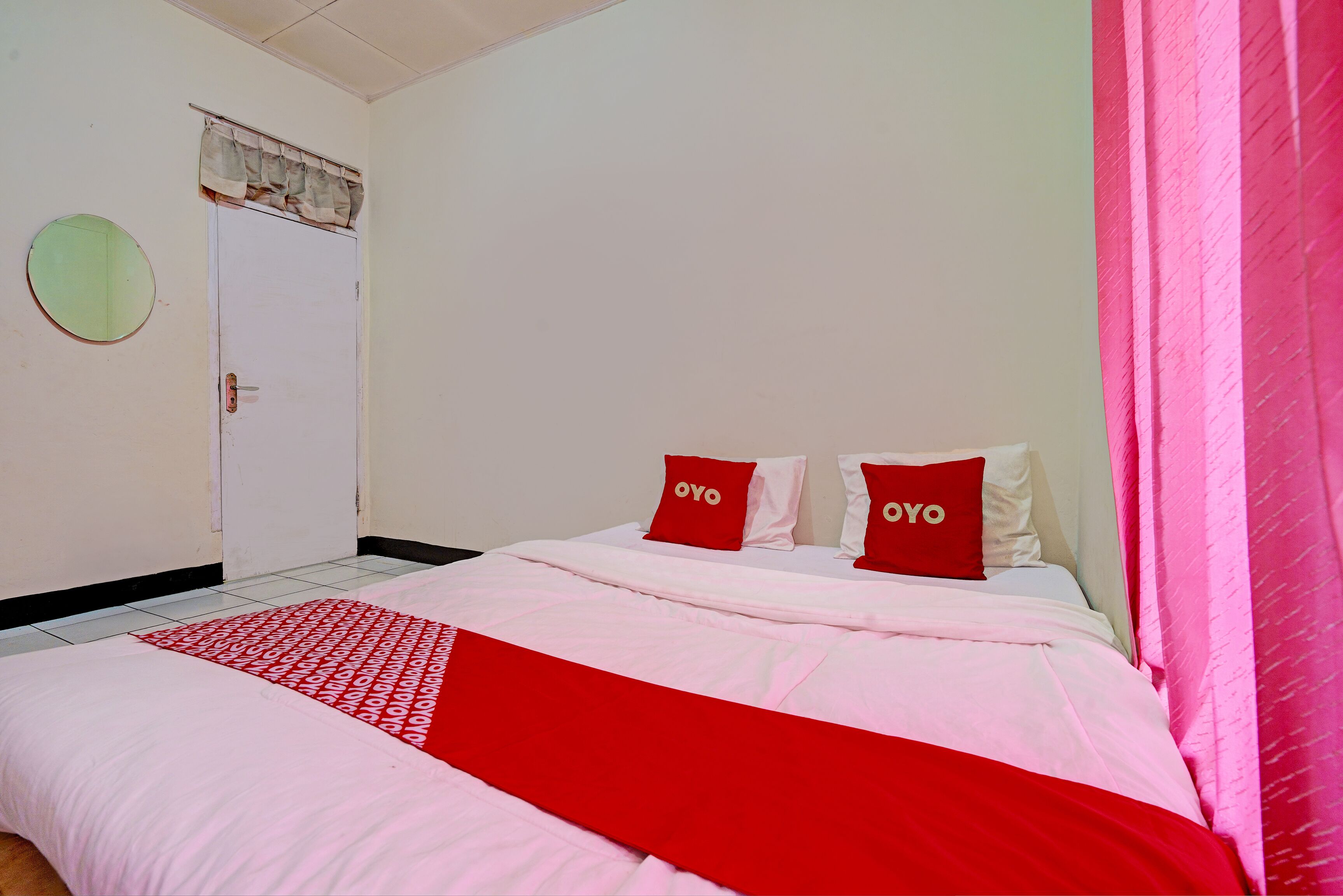 A bedroom with a bed, two pillows with "OYO" text, a red and white bedspread, a round mirror, and a white door.