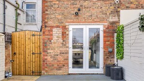 A brick house with a wooden gate, a white door, and a small garden area with a black trash bin and a potted plant. - Super Comfortable & High Quality (Cleethorpes)