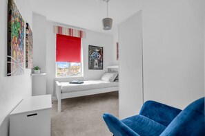 Quarto moderno com cama, uma cadeira azul, janela com persiana vermelha e vista para a cidade.
