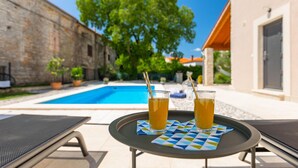 A poolside area with two glasses of orange beverage on a table, a pool, and a building with a stone facade. - Awesome home in Marcana (Marcana)