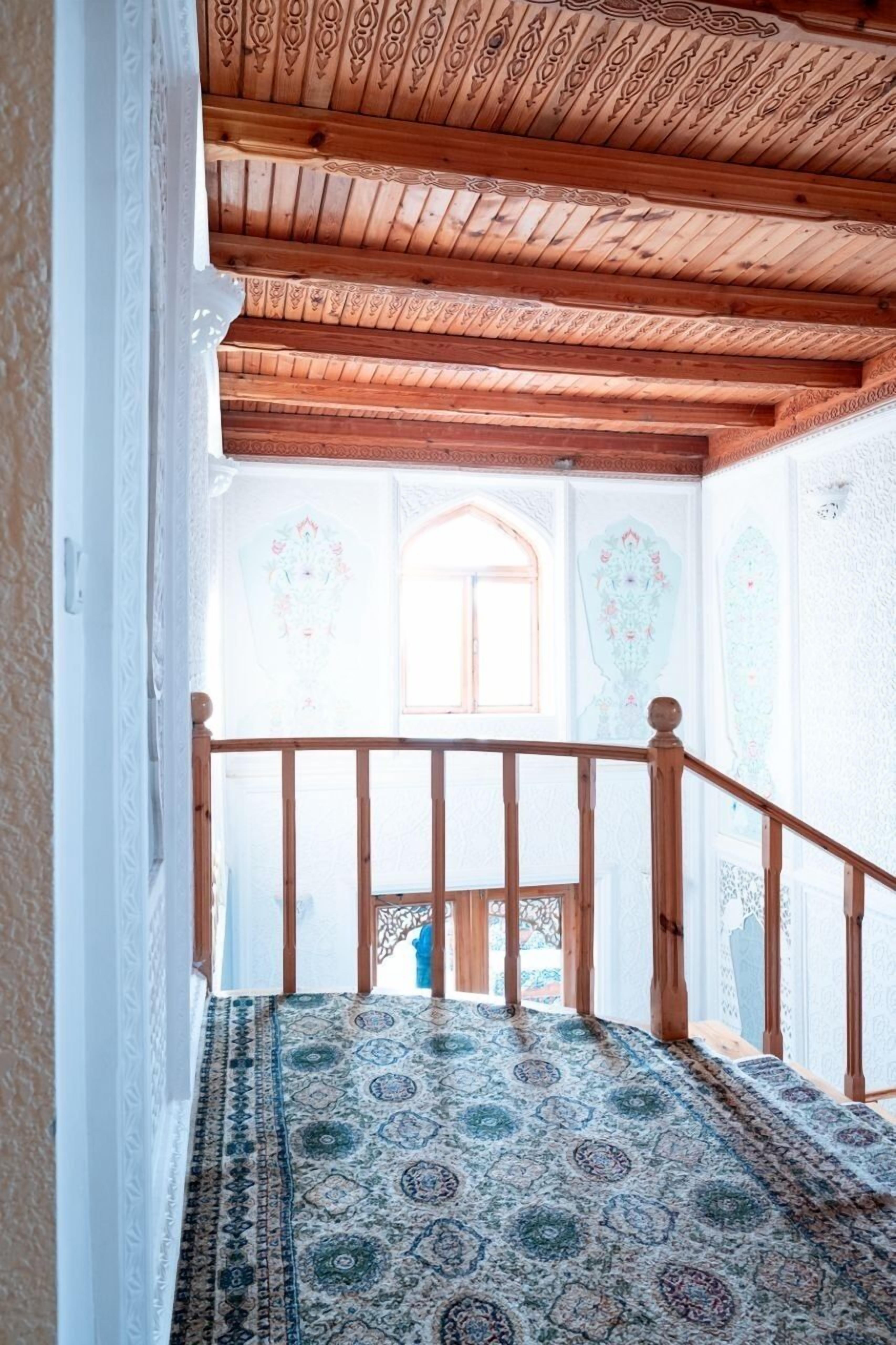 Un escalier recouvert d’une moquette à motifs mène à une pièce dotée d’un plafond en bois et d’une fenêtre ornée d’un motif décoratif.