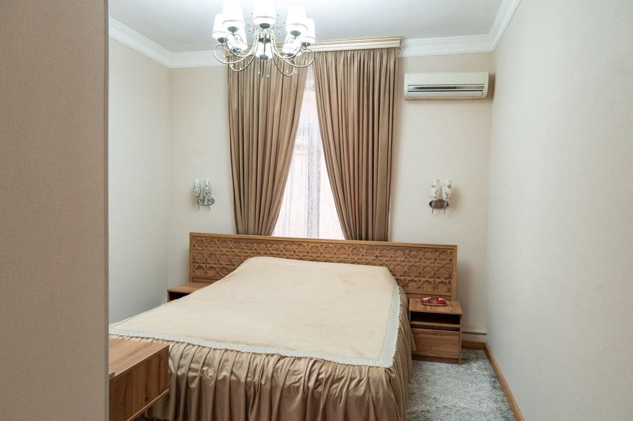 Une chambre à coucher avec un grand lit, un lustre, des rideaux, une tête de lit en bois, des tables de chevet et un appareil de climatisation.