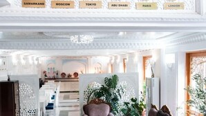 Un vestíbulo amplio con techo alto, zona de estar con sofá y sillones, pared decorativa con relojes que muestran nombres de ciudades, y vista al área de comedor.