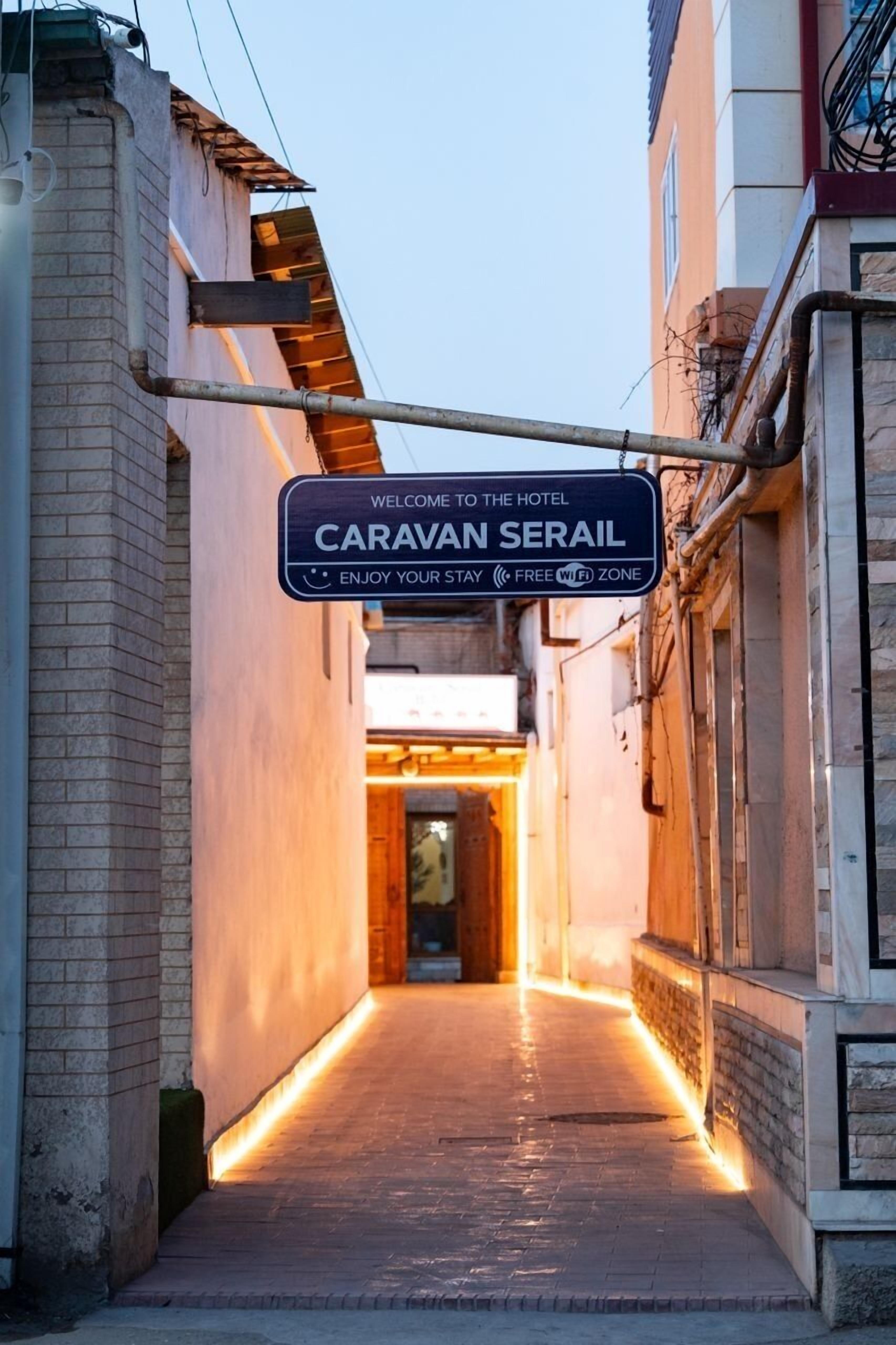 Une ruelle étroite mène à l’entrée d’un hôtel, où un panneau indique : « Bienvenue à l’hôtel Caravan Serail. Nous vous souhaitons un agréable séjour. Wi-Fi gratuit. »