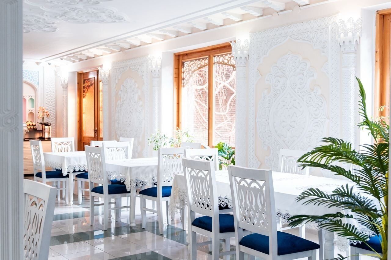 Un espace repas avec des tables et des chaises blanches, des motifs complexes sur les murs et une grande fenêtre agrémentée de volets en bois.