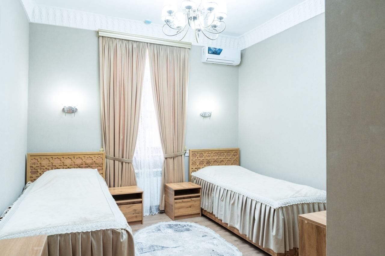 A bedroom with two single beds, a chandelier, an air conditioning unit, and a window with curtains.