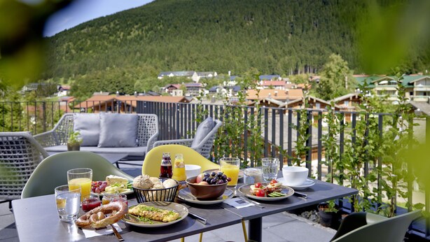 Una mesa dispuesta con alimentos para el desayuno, como pan, fruta y bebidas, con vistas a un pintoresco pueblo y colinas boscosas.
