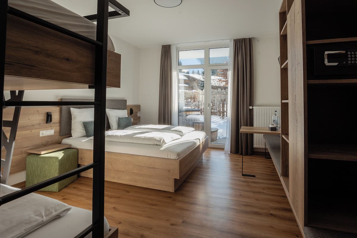 Ein modernes Schlafzimmer mit einem hölzernen Bett, einem Etagenbett, einem grünen Hocker und Blick auf eine verschneite Landschaft durch ein großes Fenster.