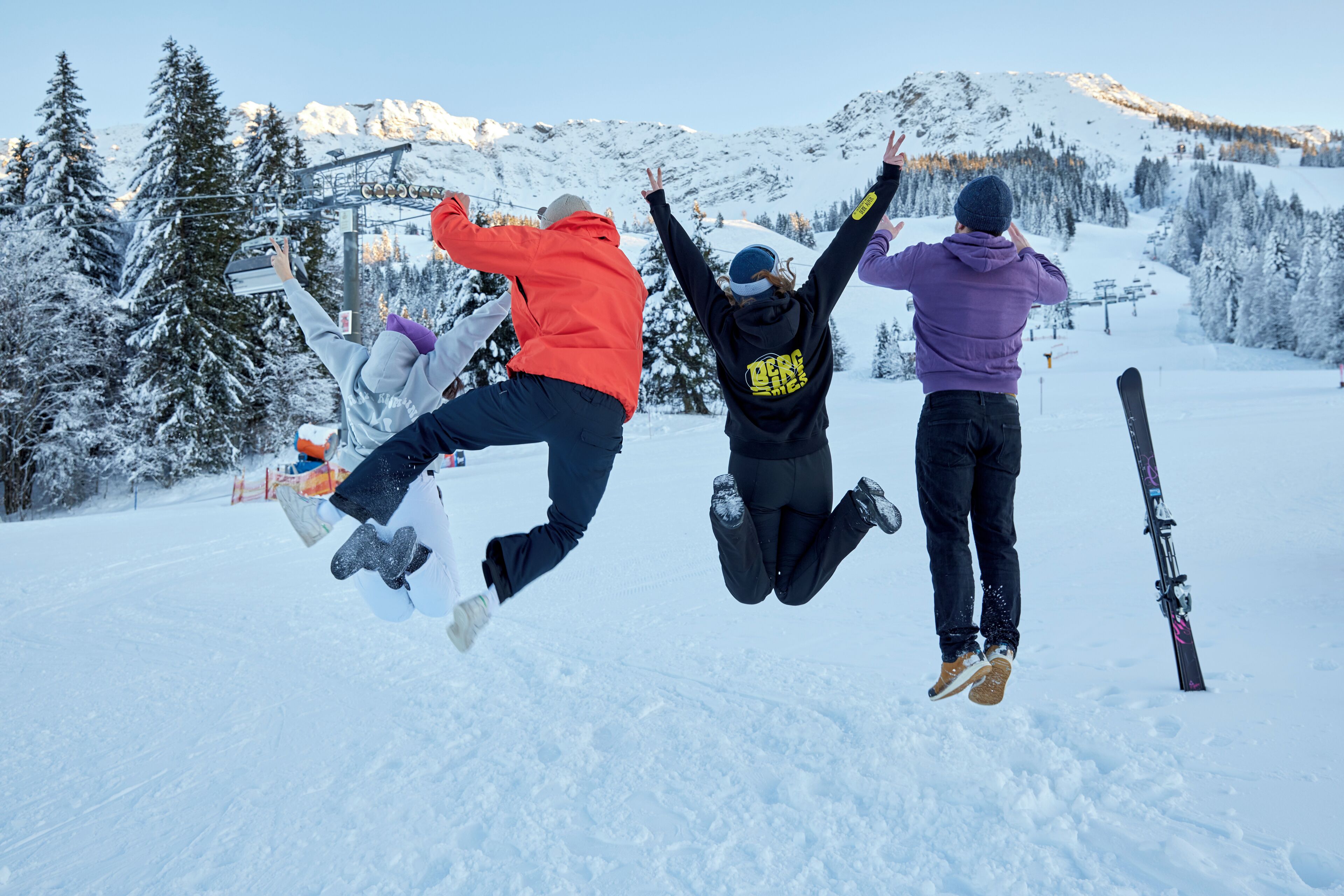 Tres personas saltando en el aire sobre una pista de esquí nevada, con equipo de esquí y un telesilla al fondo.
