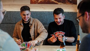 Un grupo de personas jugando cartas sobre una mesa de madera, con una botella y cartas a la vista.