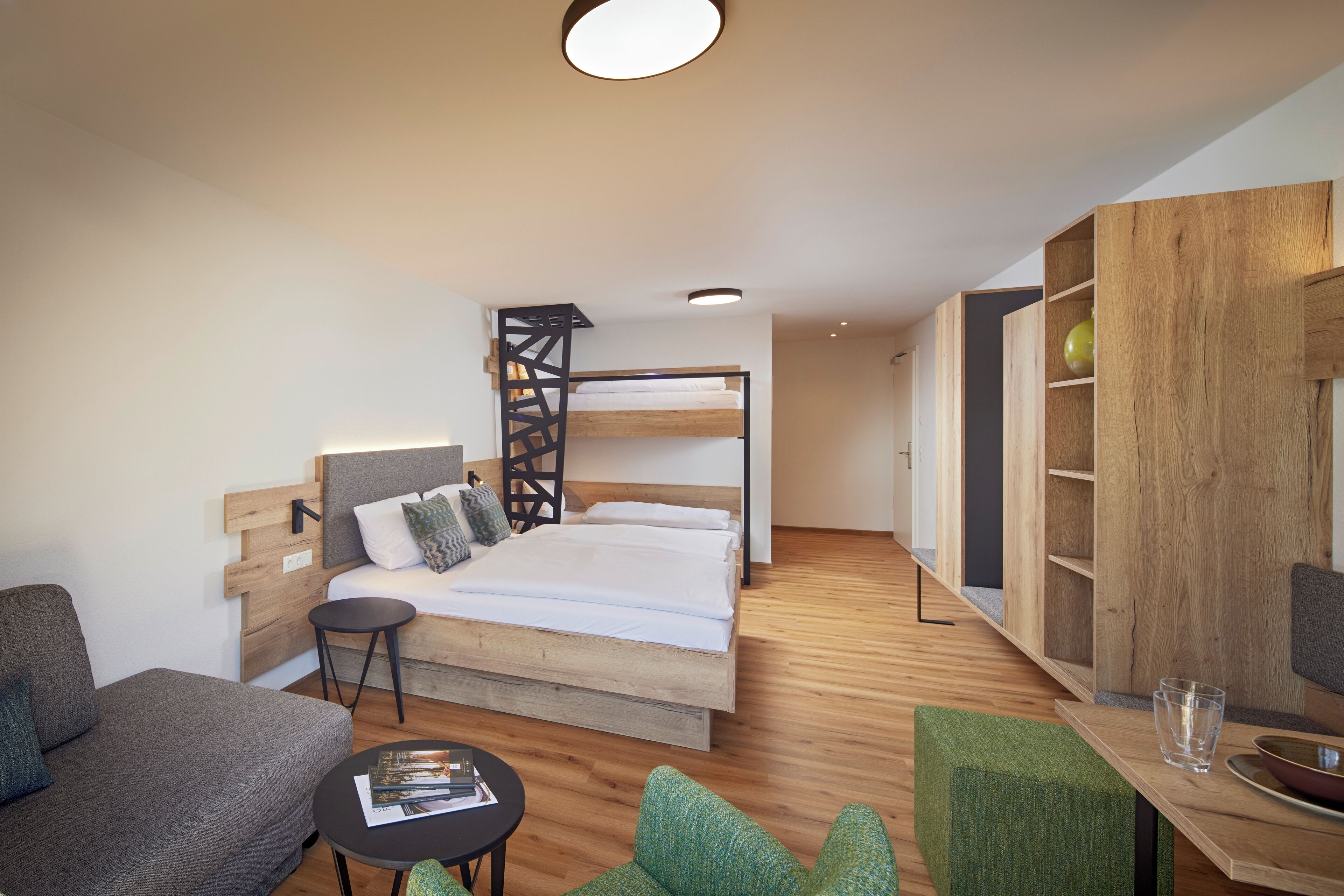 A modern hotel room with a bunk bed, a small sofa, a round table, and a wooden bookshelf.