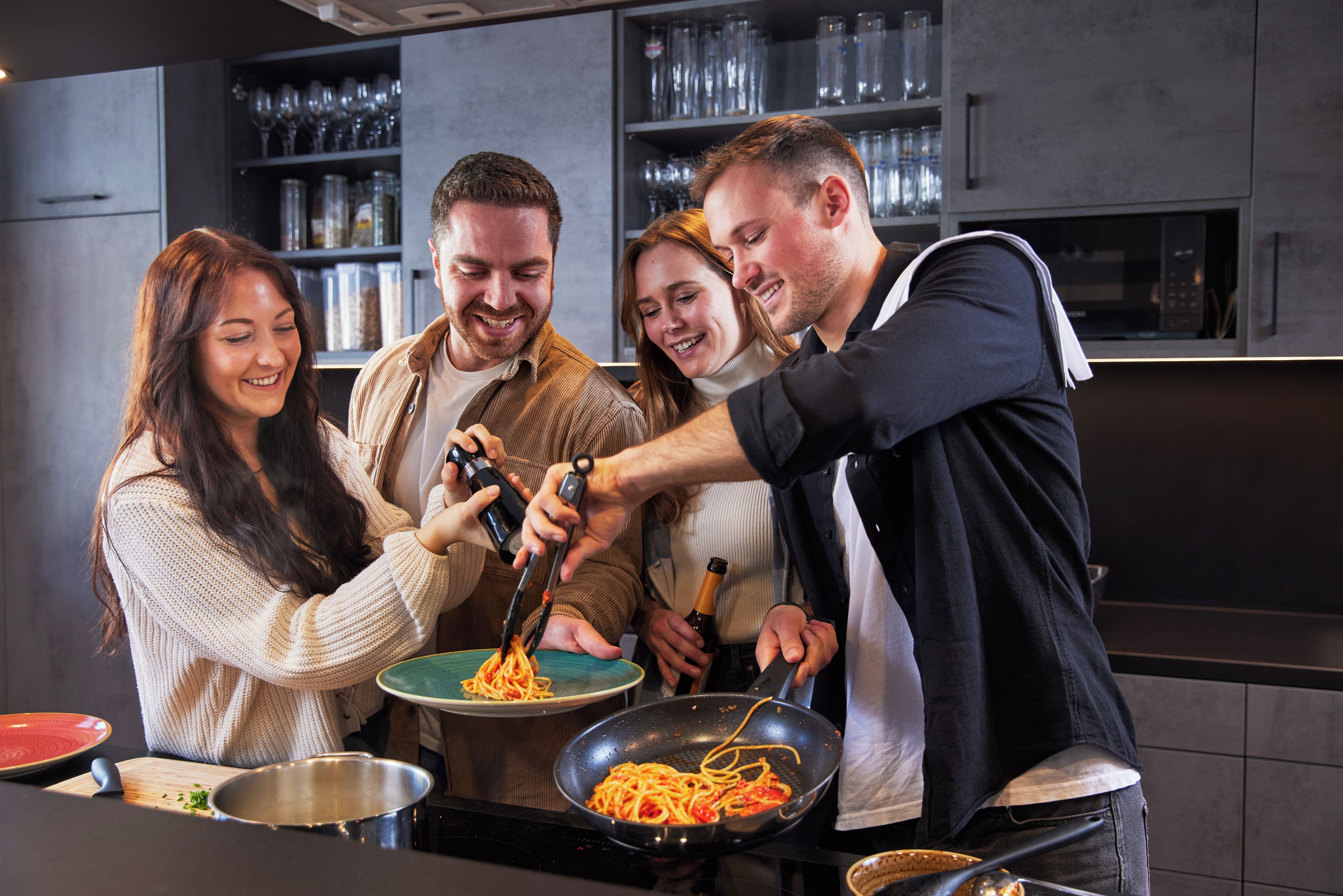 Eine Gruppe von Menschen kocht Spaghetti in einer modernen Küche mit Edelstahlgeräten und einem Weinkühlschrank im Hintergrund.