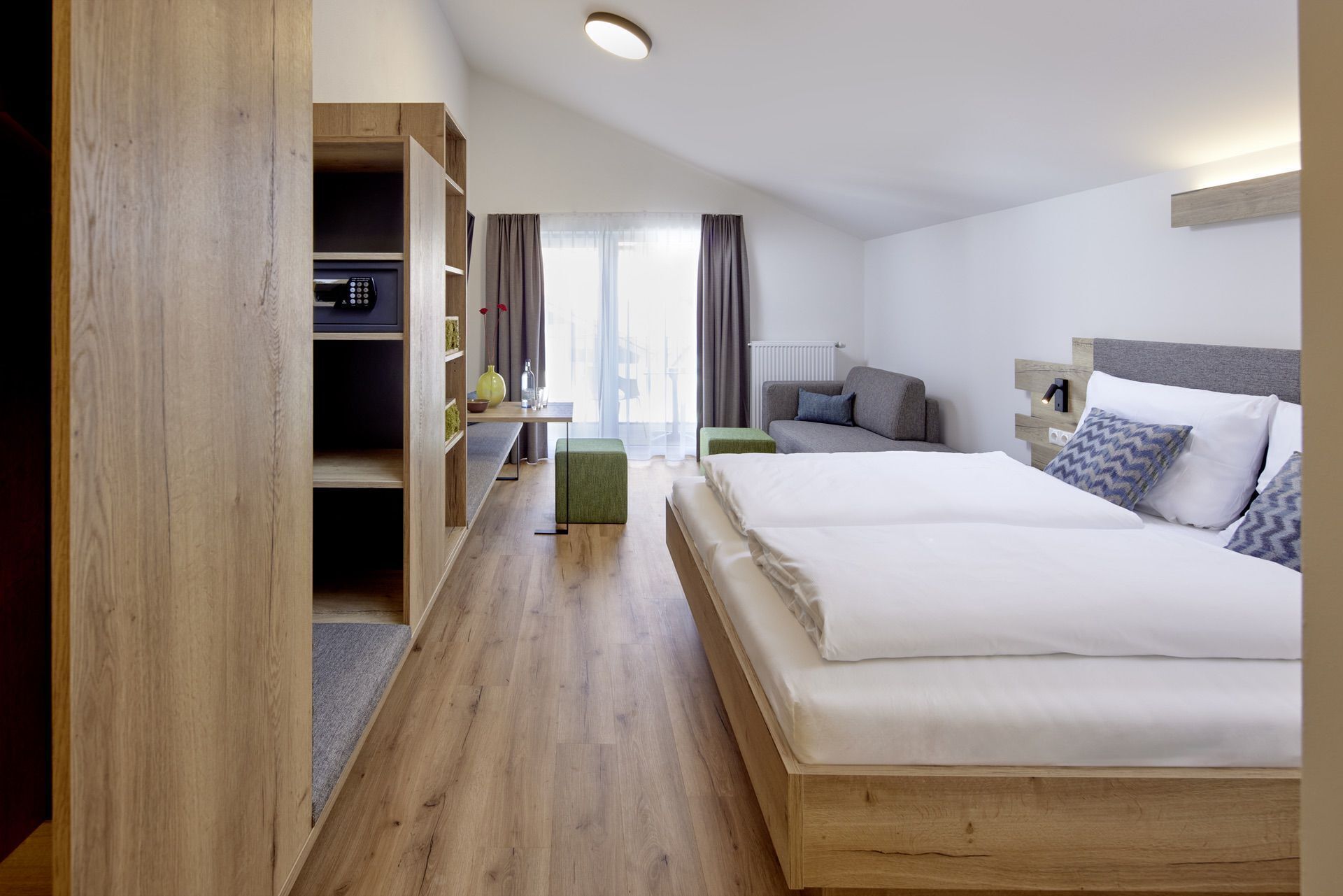 A hotel room with a wooden floor, a bed with white bedding, a grey sofa, a small table with a vase, and a wooden wardrobe.