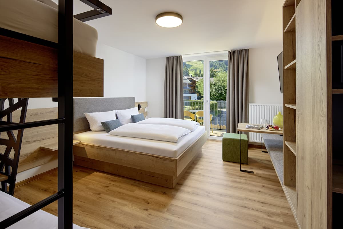Ein modernes Hotelzimmer mit einem Holzbett, einem Einzelbett, einem Schreibtisch mit Stuhl und Blick ins Freie durch große Fenster.