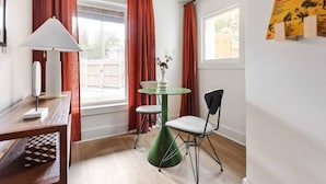 A room with a wooden floor, a round green table with two chairs, a white lamp, and a window with red curtains.