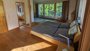 Ein Schlafzimmer mit einem großen Bett, Holzboden und Blick auf Grün hinter großen Fenstern.