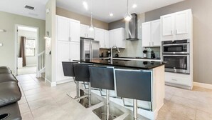 Une cuisine moderne avec des meubles blancs, un îlot central doté de tabourets de bar, des appareils en acier inoxydable et un éclairage suspendu.