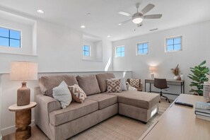 A modern living room with a sectional sofa, a wooden side table, a desk, and a ceiling fan.