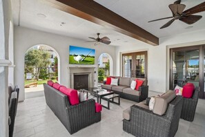 A spacious living area with wicker furniture, a fireplace, and large windows offering views of the outdoors. - Verdant Views & Amenities Near PGA West Golf (La Quinta)