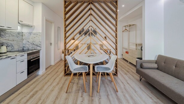 A modern kitchen with white cabinets, a marble countertop, and a built-in oven. There is a dining area with a white table and two chairs, separated by a wooden partition with a geometric design. A grey sofa is visible in the background. - Casas da Oliveira - Galega (Setúbal)