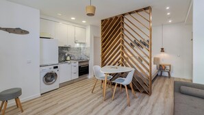 A modern kitchen with white cabinets, a built-in oven, and a washing machine. There is a dining area with a white table and chairs, and a grey sofa. - Casas da Oliveira - Galega (Setúbal)