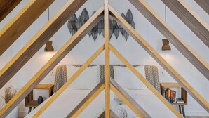 A room with a sloped ceiling, wooden beams, a bed with patterned bedding, a wall-mounted shelf with books, and a decorative wall piece. - Casas da Oliveira - Galega (Setúbal)