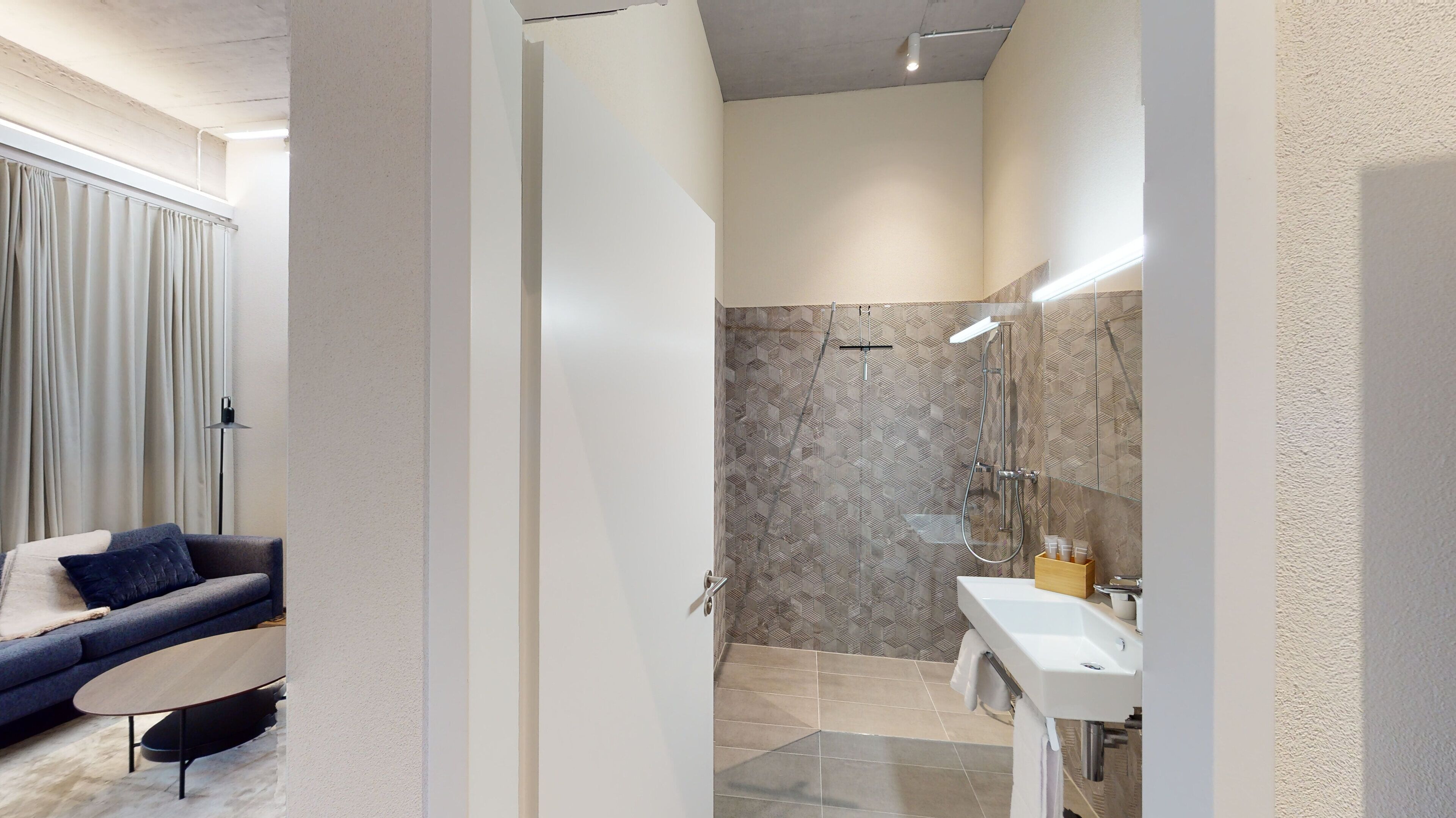 Une salle de bain moderne dotée d’une douche, d’un meuble-lavabo et offrant une vue sur un espace de vie comprenant un canapé et une lampe.