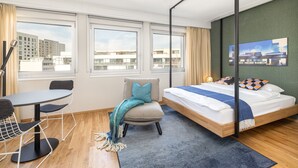 A modern hotel room with a bed, a desk, and a chair. There are large windows with a city view. - LivingTown Lofts Zurich (Zürich)