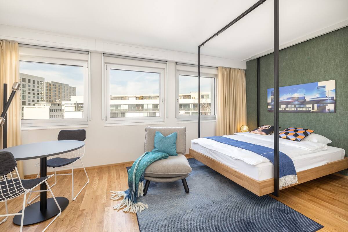 A modern hotel room with a bed, a desk, and a chair. There are large windows with a city view.