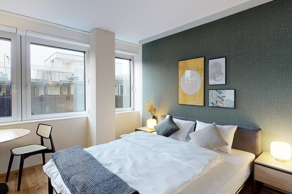 A modern hotel room with a large bed, a bedside table with a lamp, a chair, and a view of the outside through large windows.