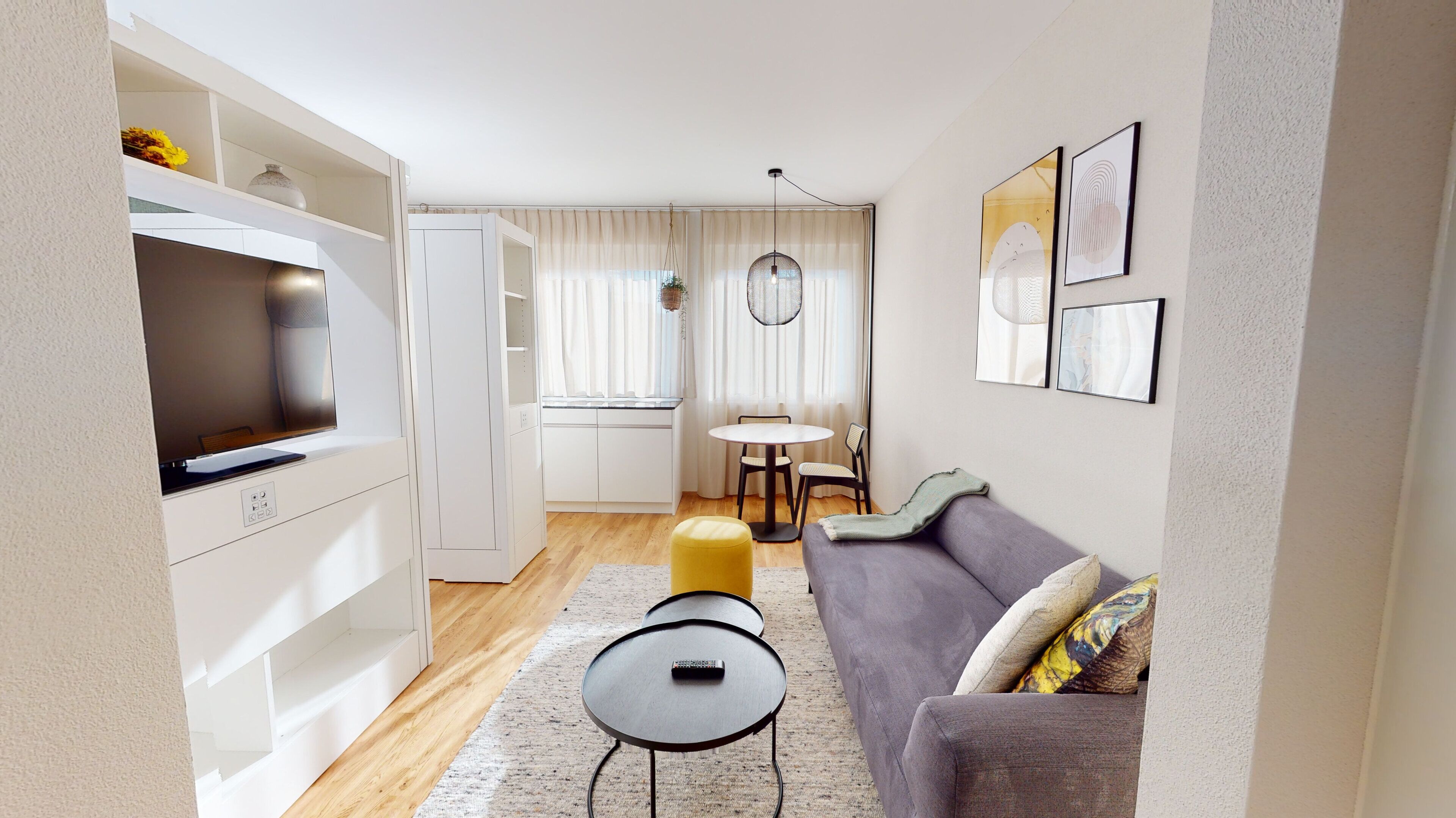 Una sala moderna con un sofá gris, una mesita redonda y un televisor. Se observa un comedor con una mesa y sillas, y una cocina con gabinetes blancos.
