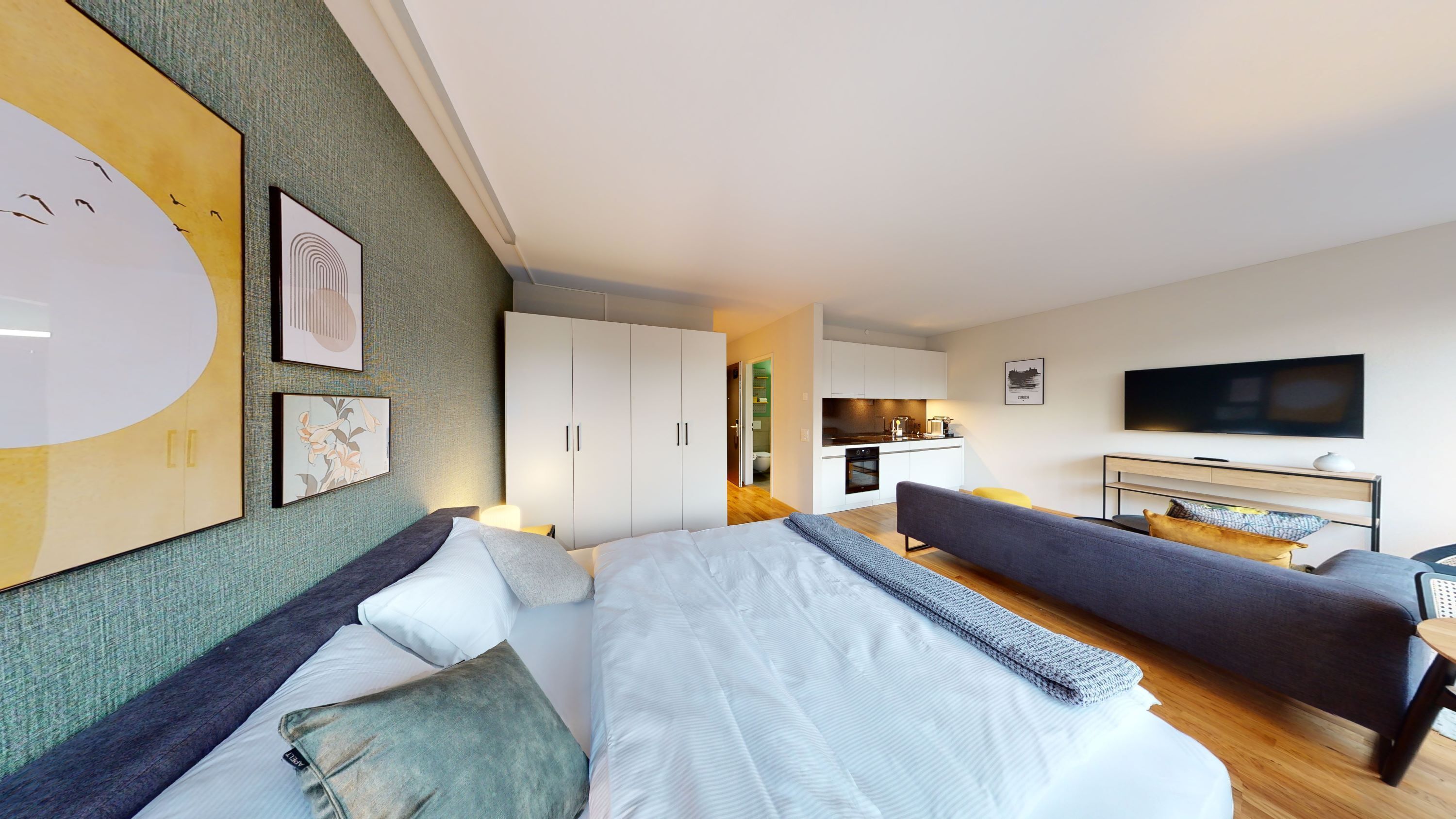 A modern hotel room with a large bed, a sofa, a TV, and a kitchenette in the background.