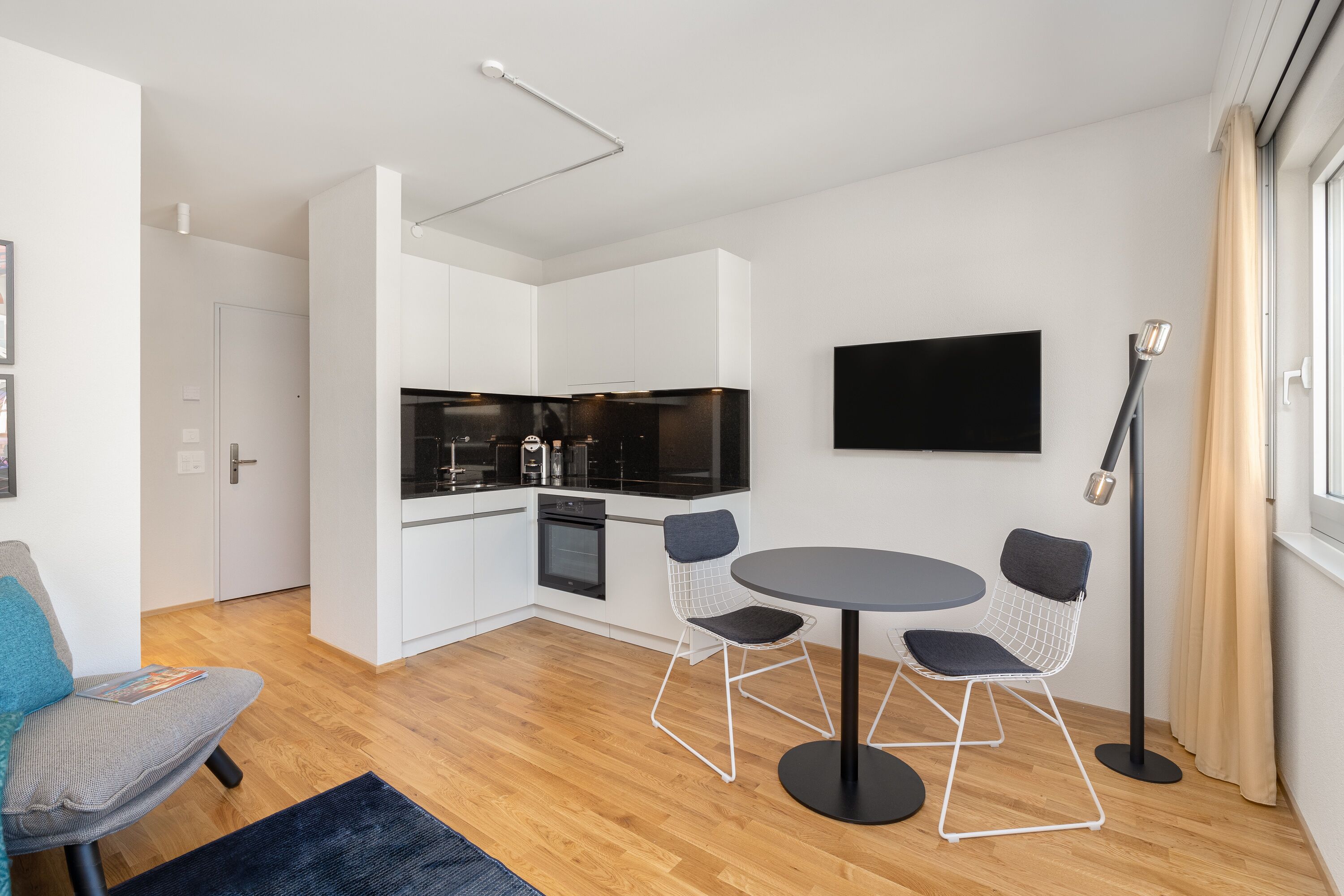 A modern kitchen with white cabinets, a black countertop, and a dining area with a round table and wireframe chairs.