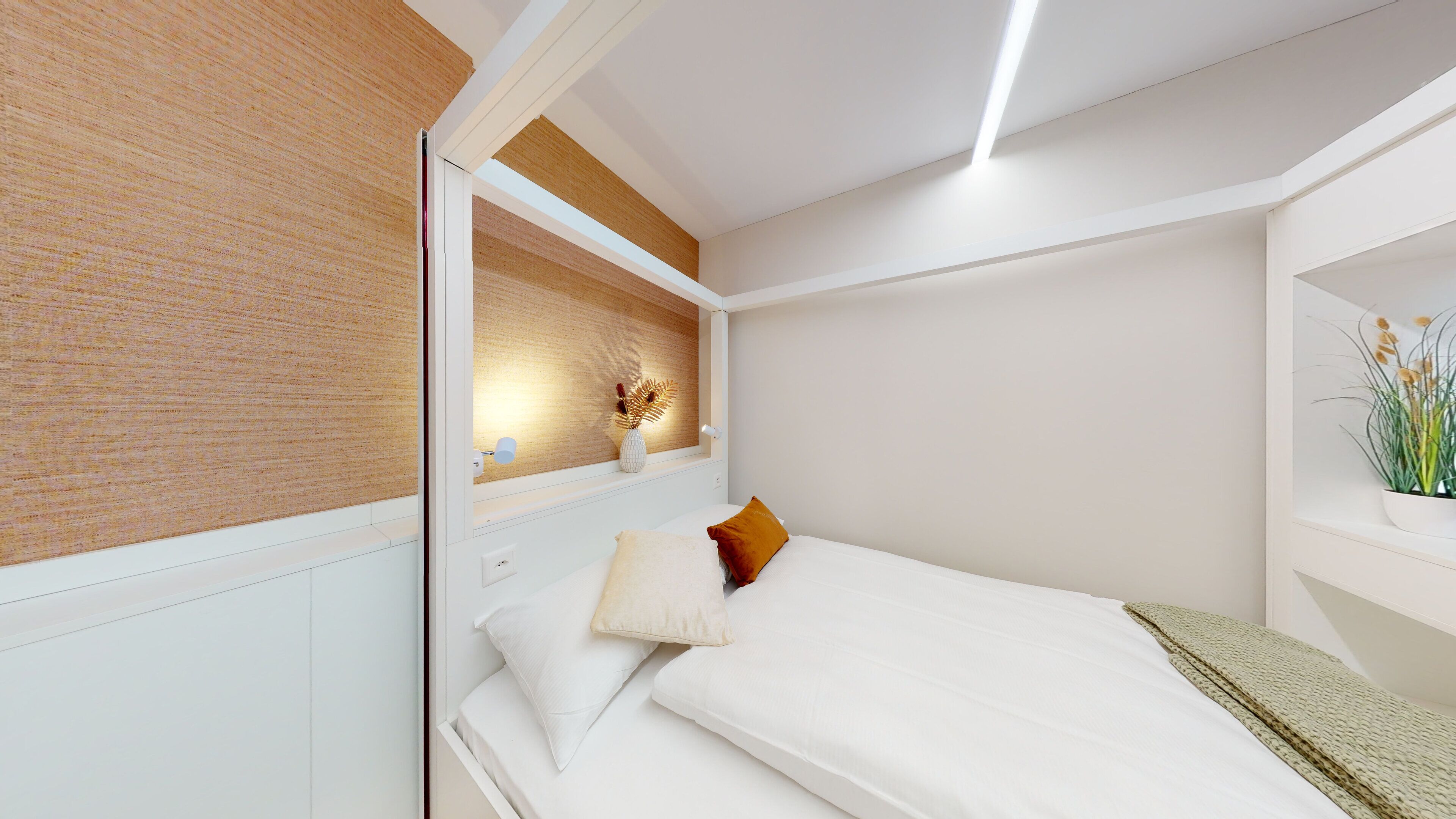 A modern bedroom with a bed, white bedding, a wooden headboard, a bedside table with a vase, and a wall-mounted shelf with a plant.