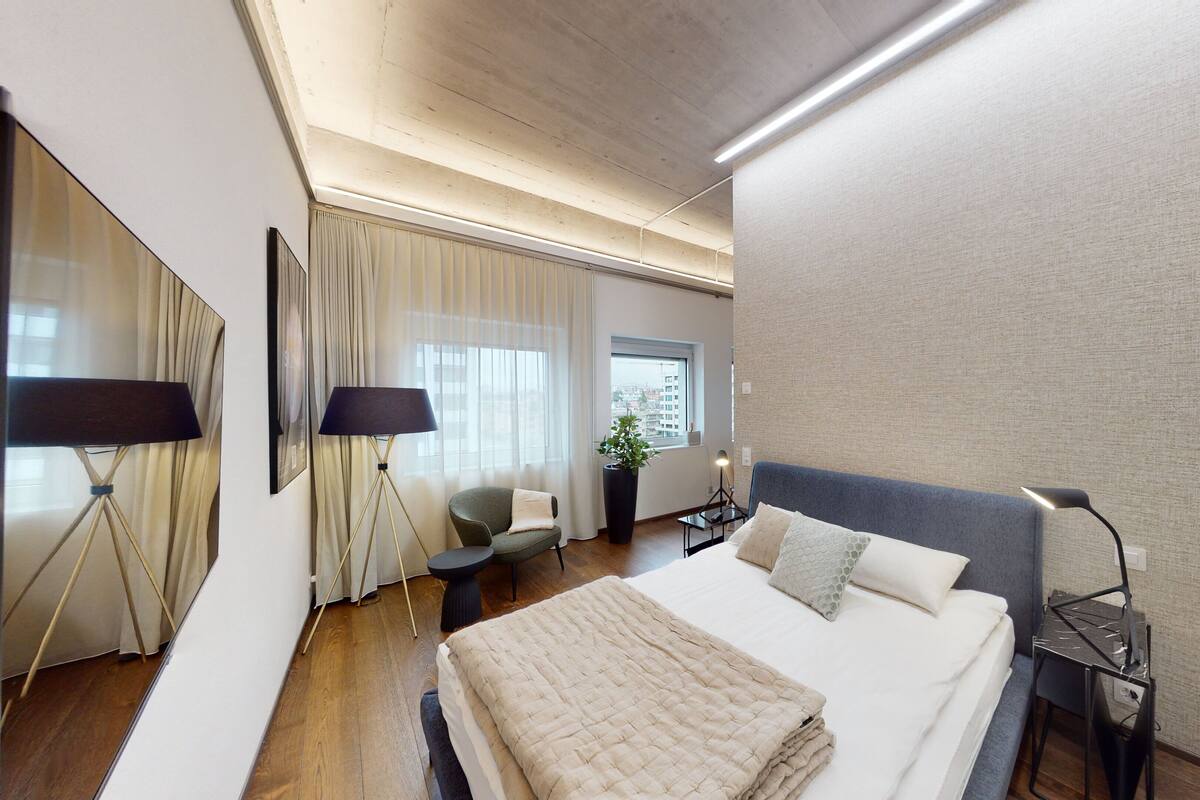 A modern hotel room with a large bed, a desk, a chair, and a window with curtains.