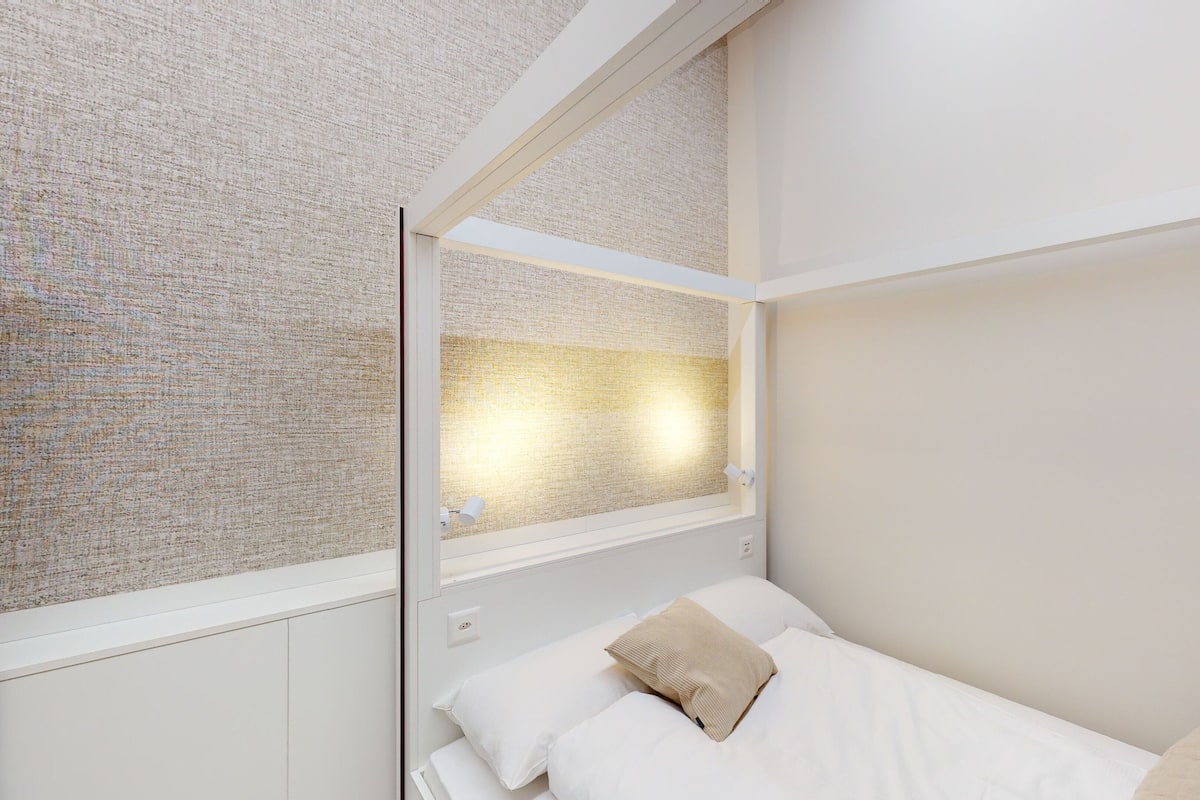 A bedroom with a bunk bed, a window with a textured wall, and a bedside lamp.