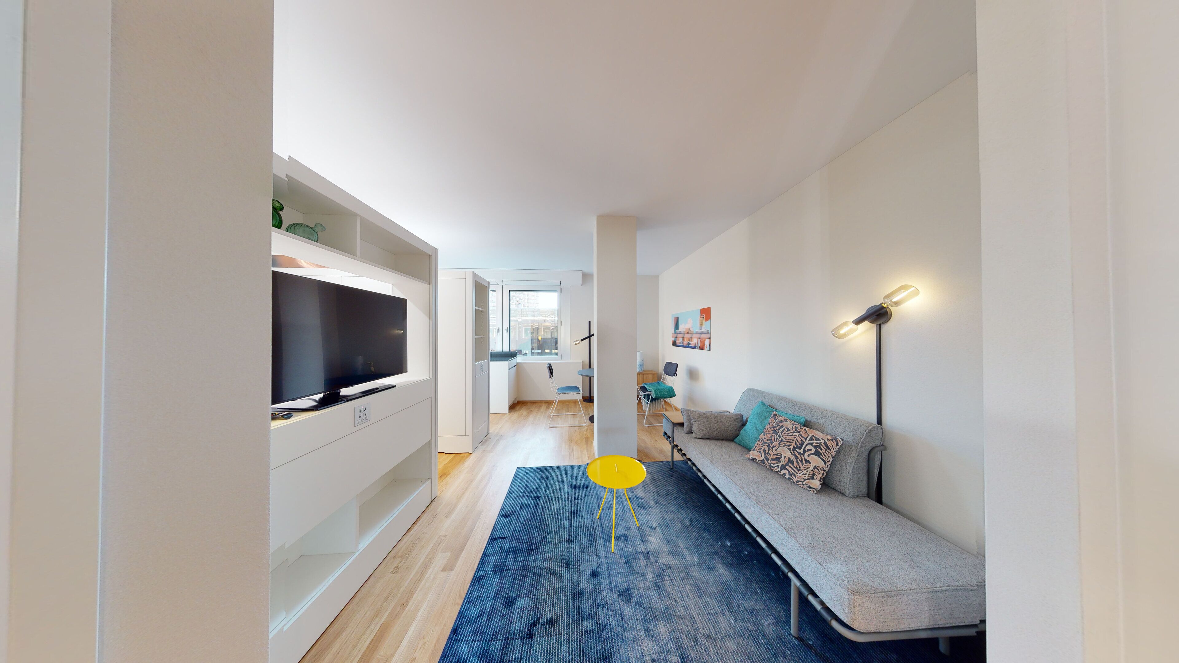 A modern apartment with a TV in the living area, a sofa with cushions, a blue rug, and a lamp.