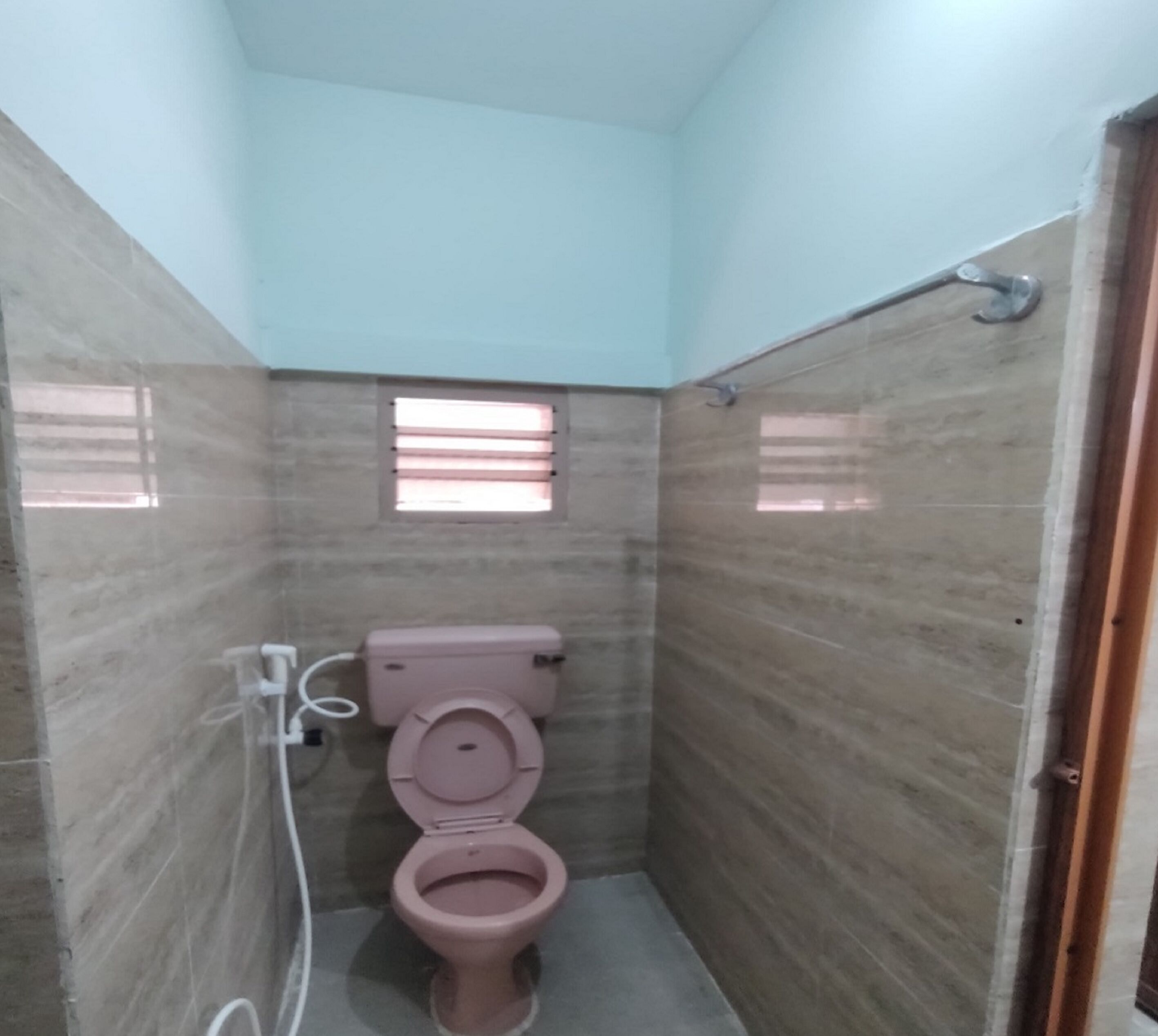 Een kleine, smalle badkamer met een roze toilet, een raam met horizontale zonwering en betegelde muren.