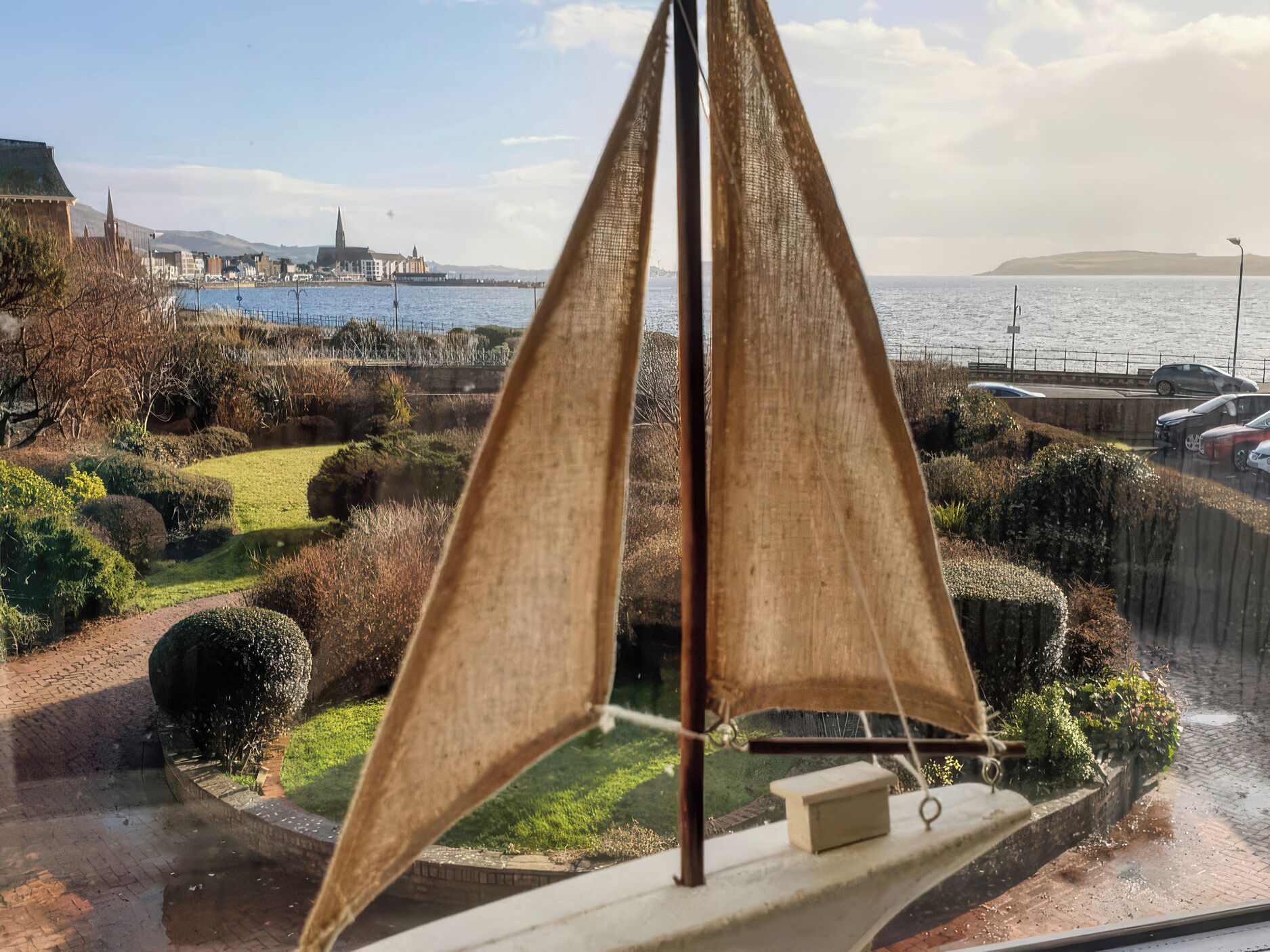 Un velero con mástil y vela sencillos, amarrado en una orilla con vista a un paisaje urbano de fondo.