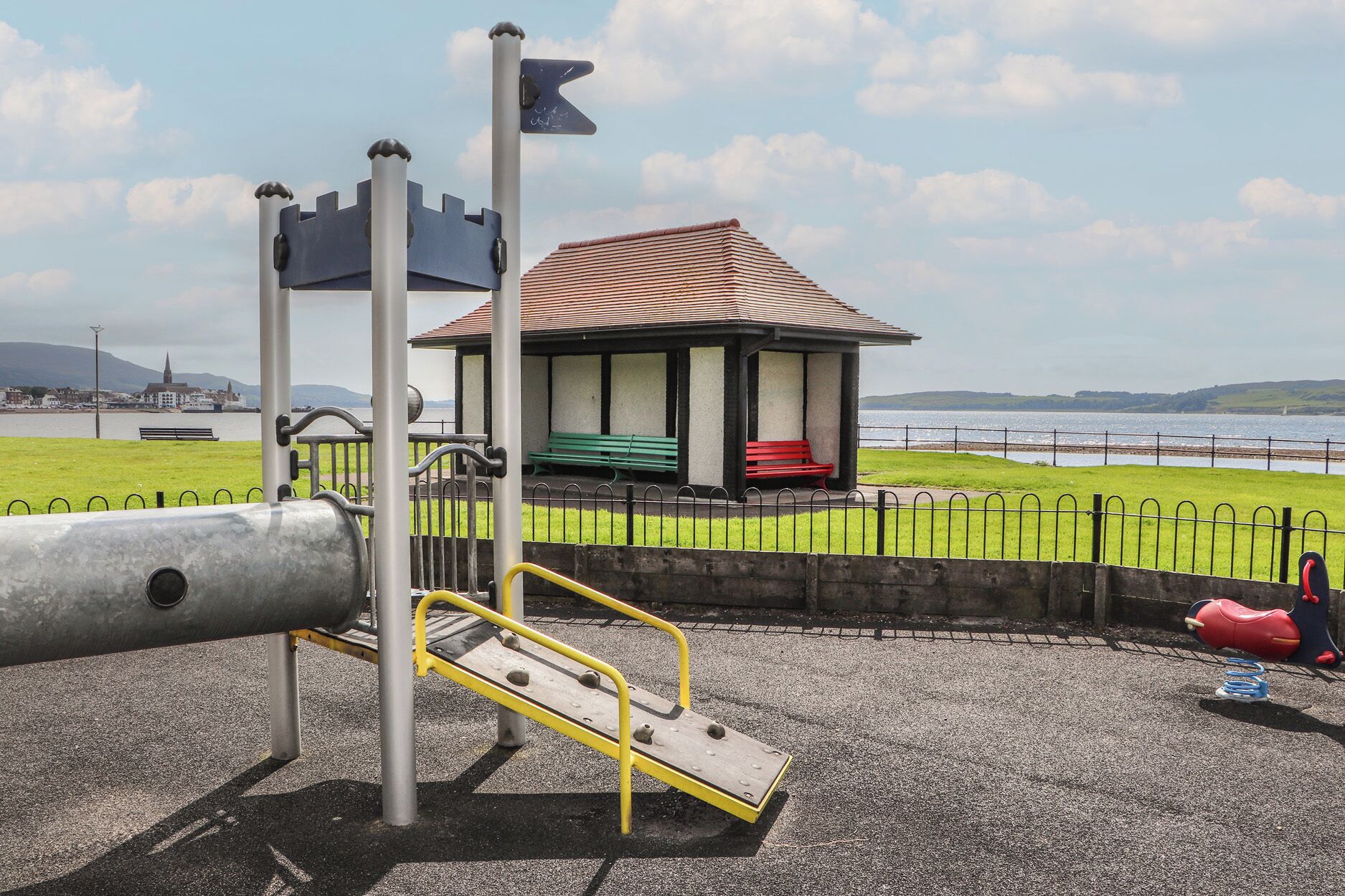 A playground with a slide and climbing structure, benches, and a small pavilion by a waterfront.