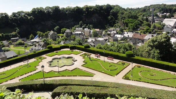 Un jardín formal con diseños geométricos y una fuente central, rodeado de setos cuidadosamente recortados y senderos.