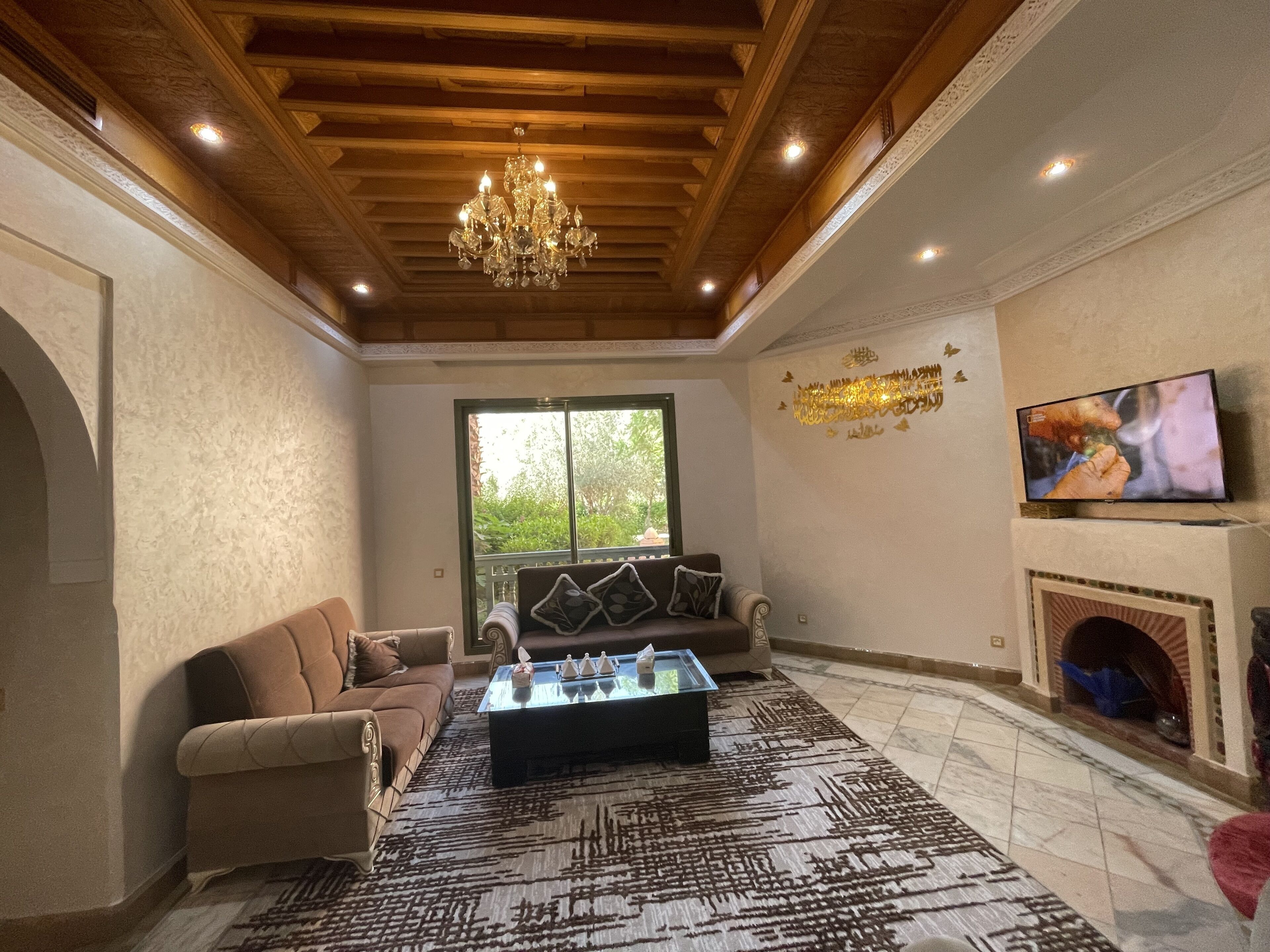 A spacious living room with a wooden ceiling, a chandelier, a sofa set, a coffee table, a fireplace, and a television.
