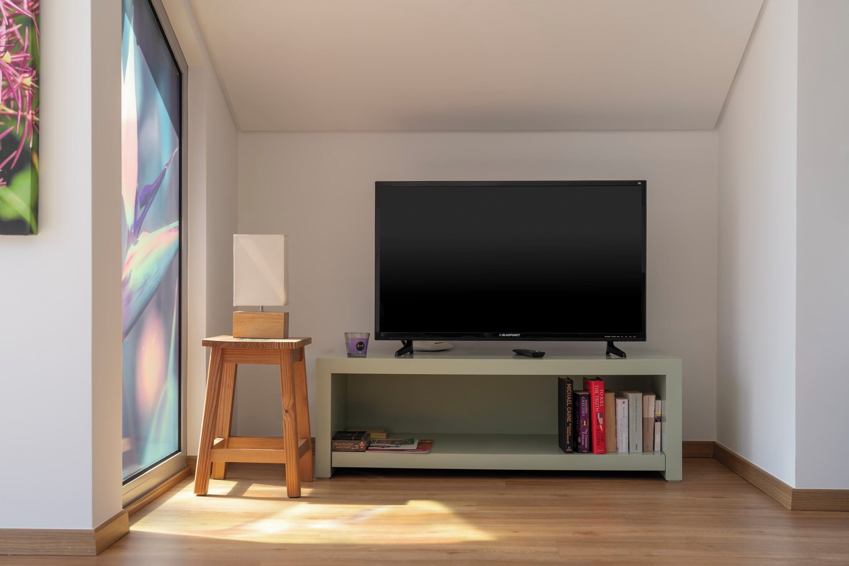A modern living room with a flat-screen TV on a stand, a small wooden stool, and a bookshelf with books.
