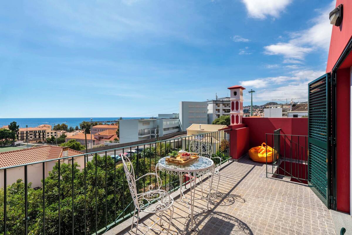 Een balkon met een tafel en stoelen, uitzicht op zee en gebouwen op de achtergrond.