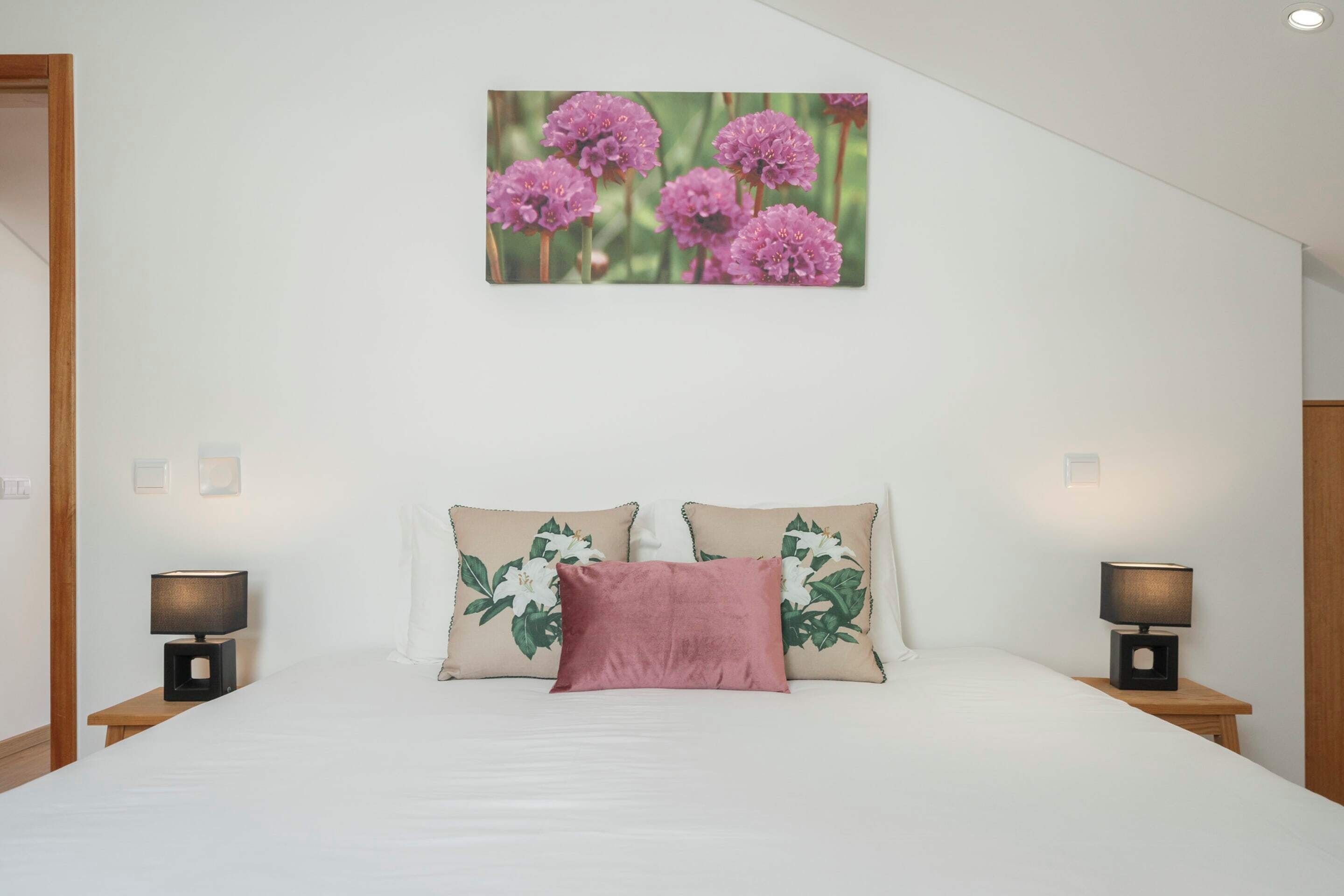 A bedroom with a bed, two nightstands, a wall-mounted lamp, and a floral print pillow.
